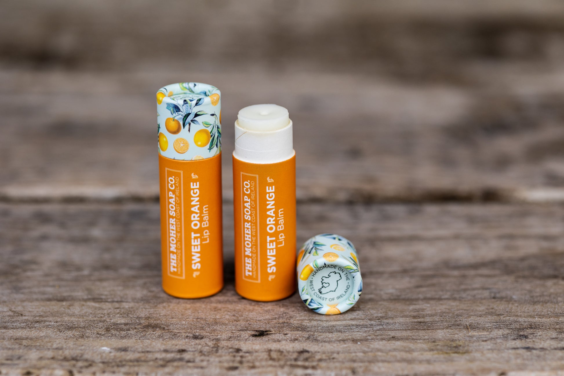 Two Sweet Orange Lip Balms by The Moher Soap Co, one opened to reveal balm, with a citrus patterned cap nearby.