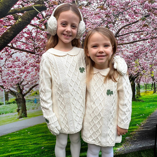 Two girls wearing matching cream Aran knit dresses with shamrock patches posing outdoors among blooming trees.