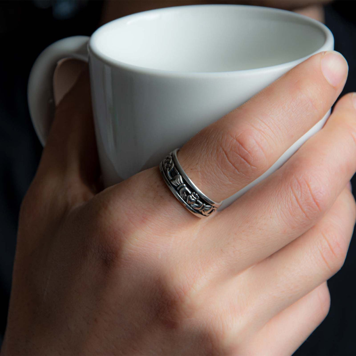 Hand wearing hallmarked sterling silver oxidised Claddagh ring on the ring finger while holding a mug.