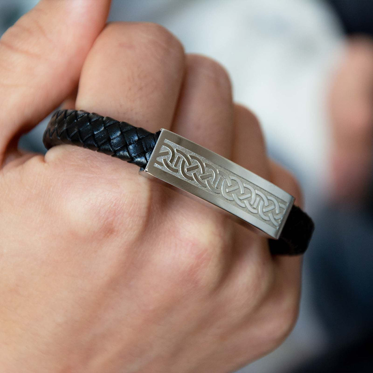 Close up of black braided leather bracelet showing a wide steel plate engraved with a continuous Celtic knot pattern