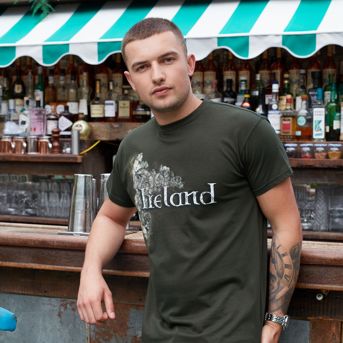 Young man with tattoo leaning on bar counter with bottles behind him