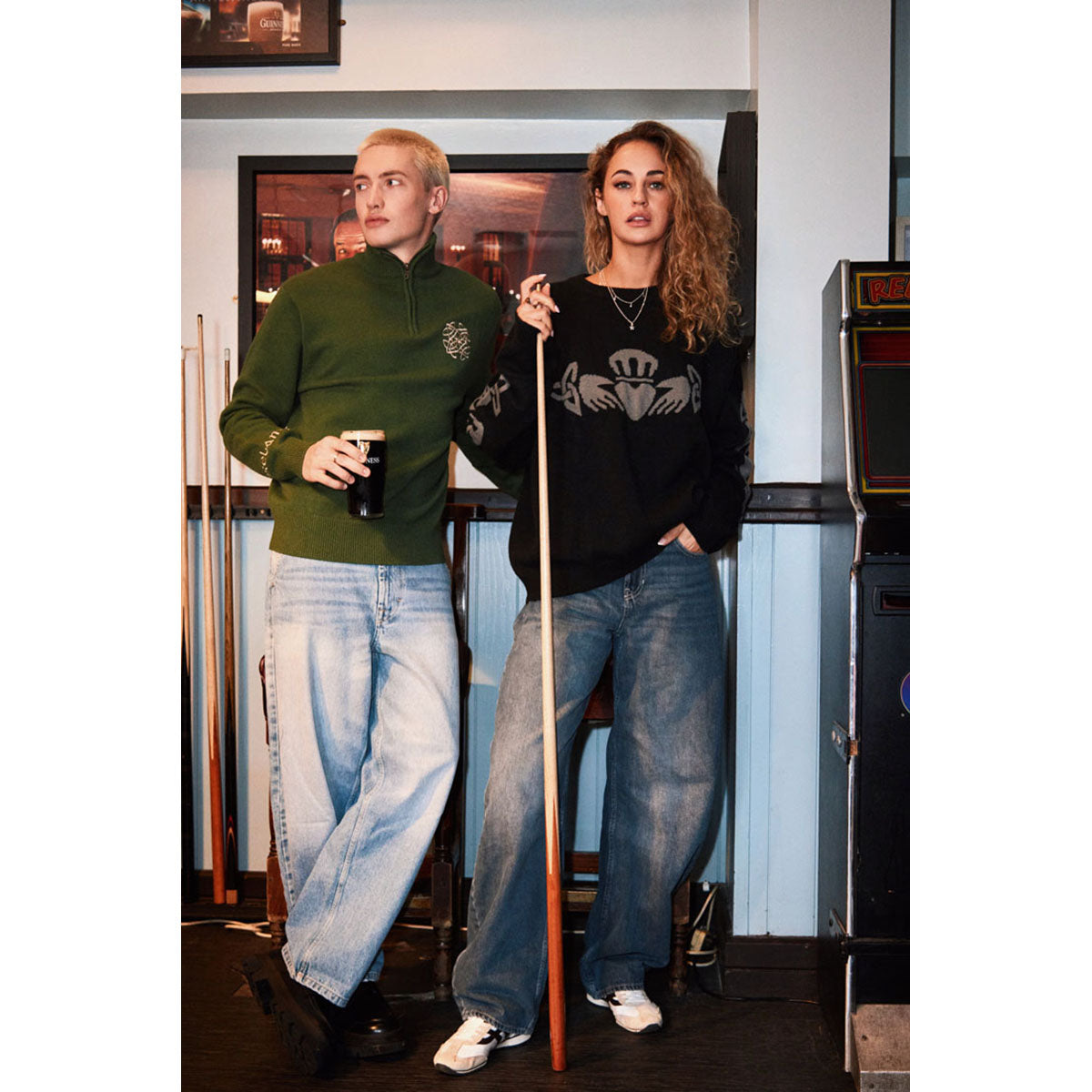 Two models wear Claddagh crew neck sweaters in a casual bar setting, showcasing a black and gray crest on the chest.