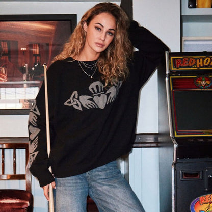 Woman wearing a black crew neck Claddagh sweater with a grey motif across the chest posing beside an arcade cabinet.