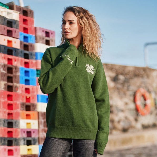 Model wearing sage green Celtic Knot 1/4 zip sweater with white chest logo outdoors near colorful pallets.