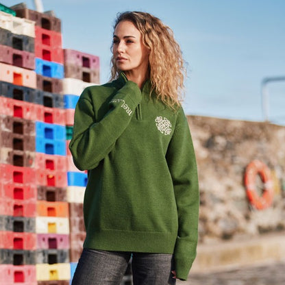 Model wearing sage green Celtic Knot 1/4 zip sweater with white chest logo outdoors near colorful pallets.
