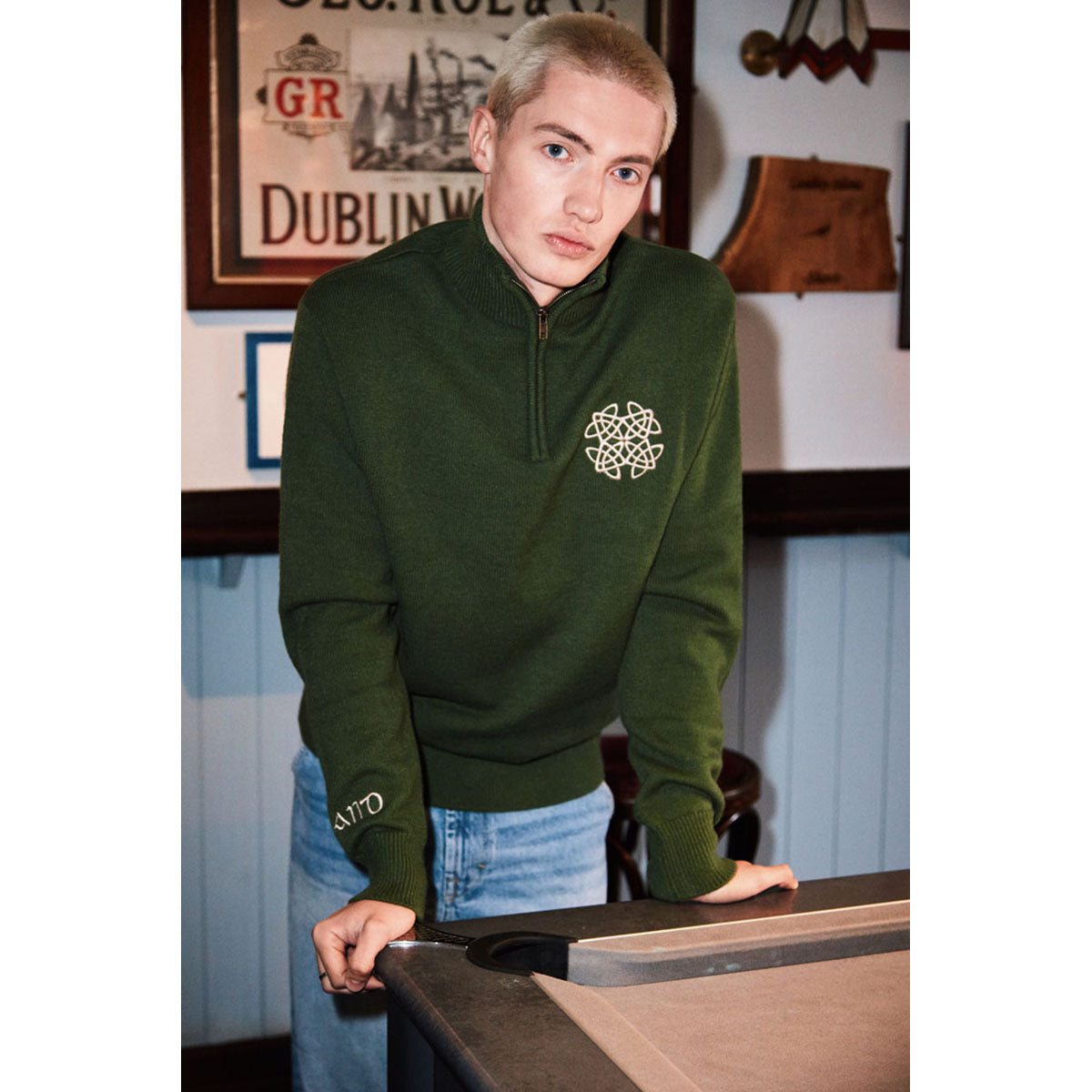 Male model wearing sage green Celtic Knot 1/4 zip sweater with chest logo indoors.