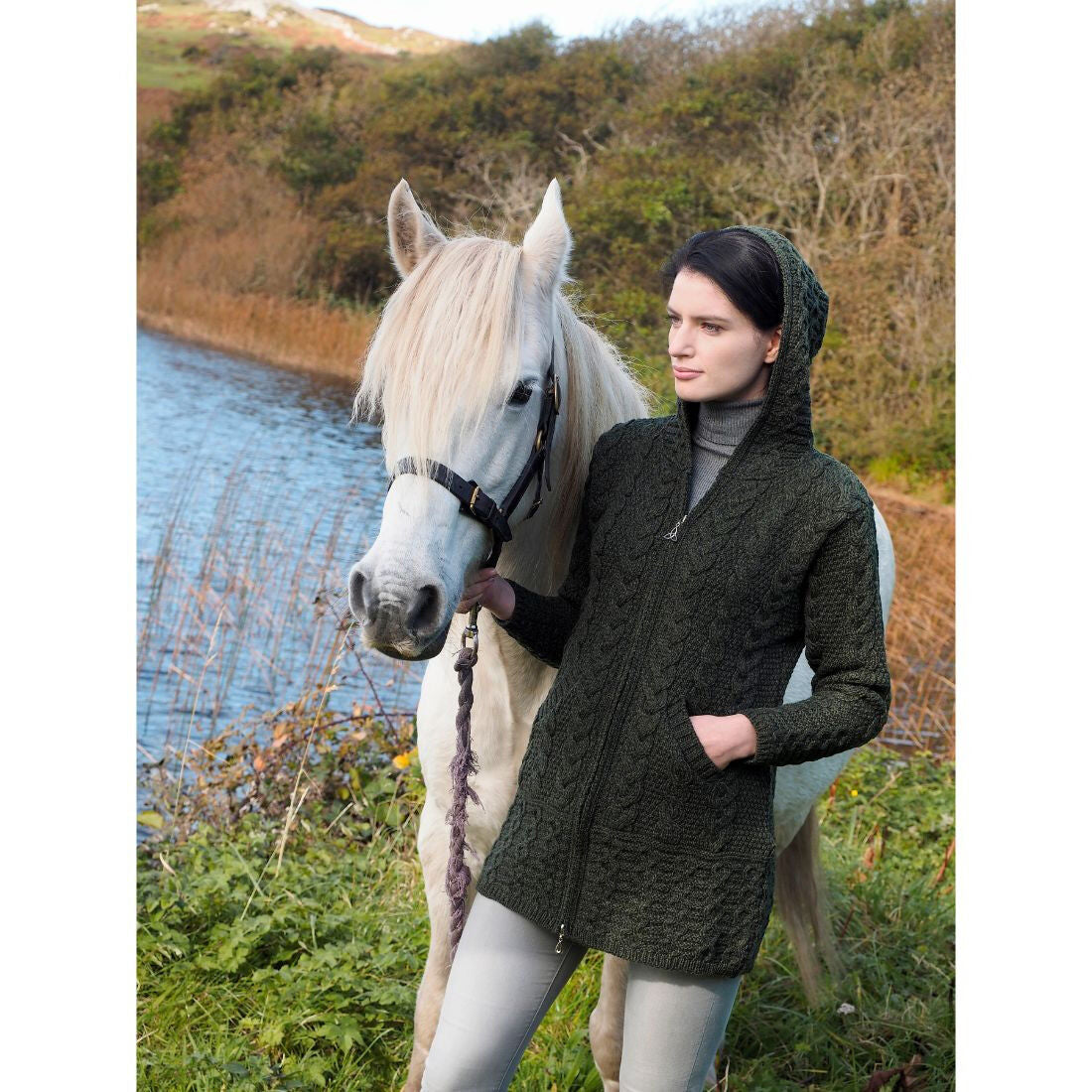 Woman in dark hooded sweater standing beside white horse near water