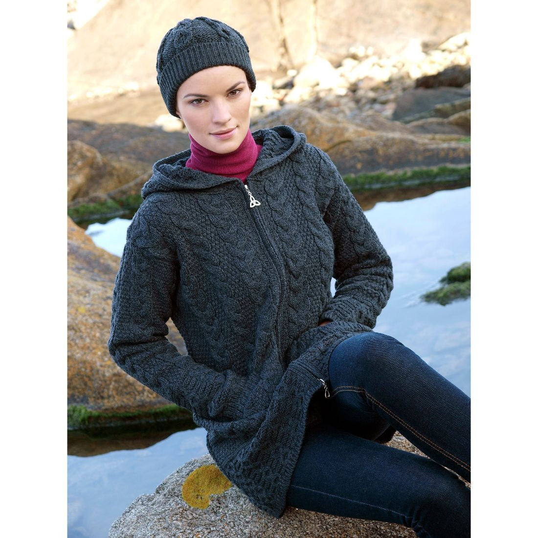 Woman wearing dark knitted hat and sweater sitting on rock near water