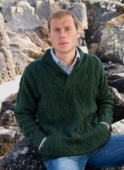 Male model wearing a dark green shawl collar Aran sweater standing among rugged rocks at the beach