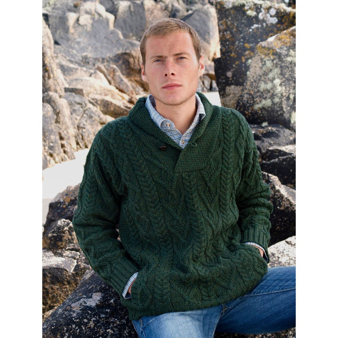 Male model wearing a dark green shawl collar Aran sweater standing among rugged rocks at the beach