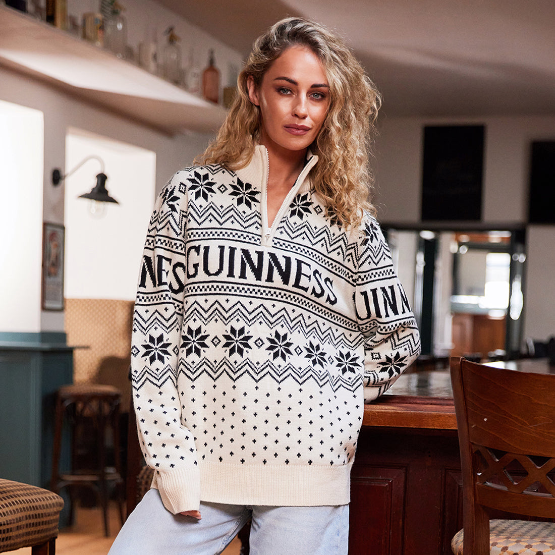 Woman wearing a cream Guinness festive knit sweater with a high zip collar and bold geometric pattern, standing by a bar.