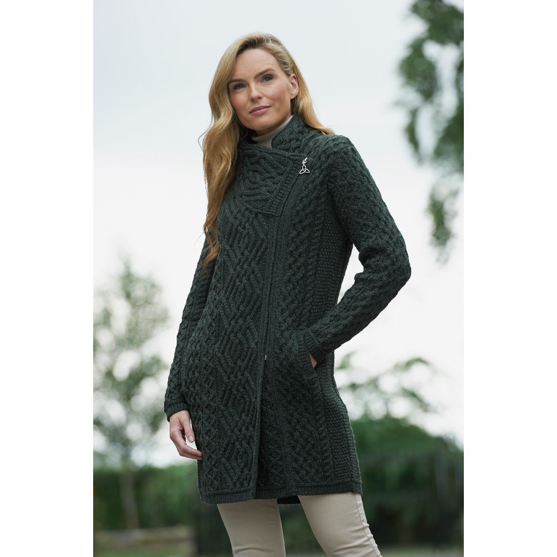 Model wearing a long dark green Aran cable knit coat with a side zip outdoors